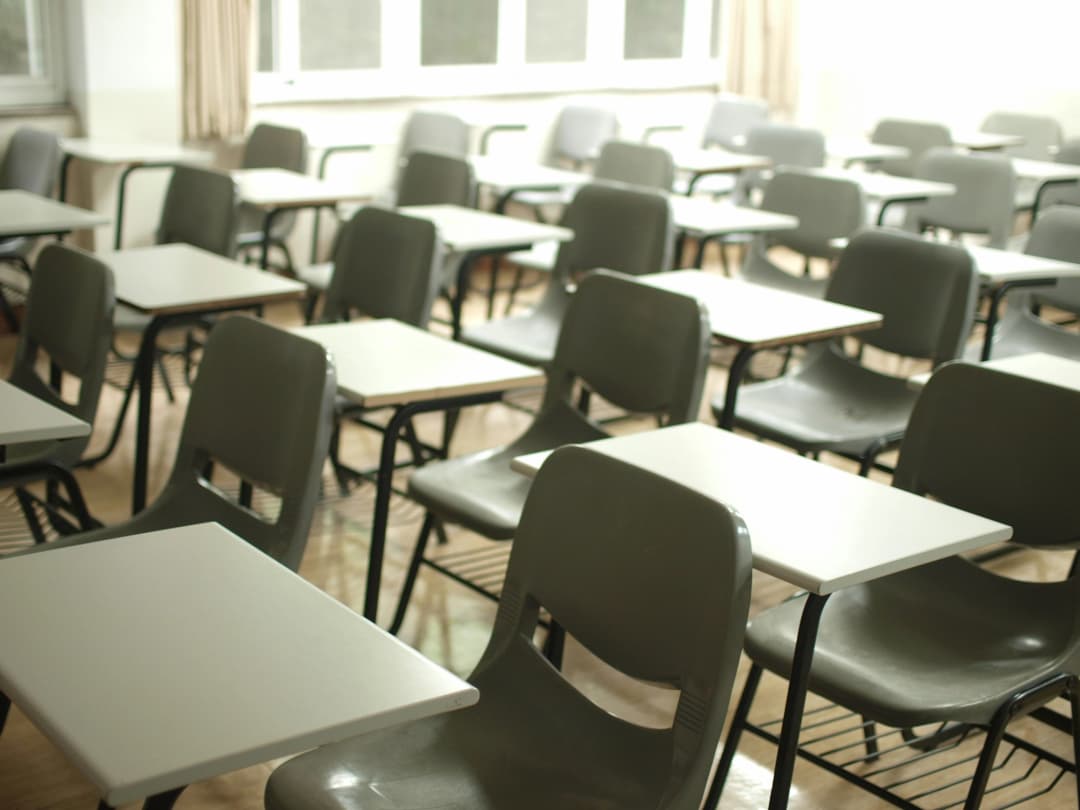 Empty classroom filled with rows of gray plastic chairs attached to white desks, lit by natural light from large windows, evoking themes of education, learning environments, or standardized testing.
