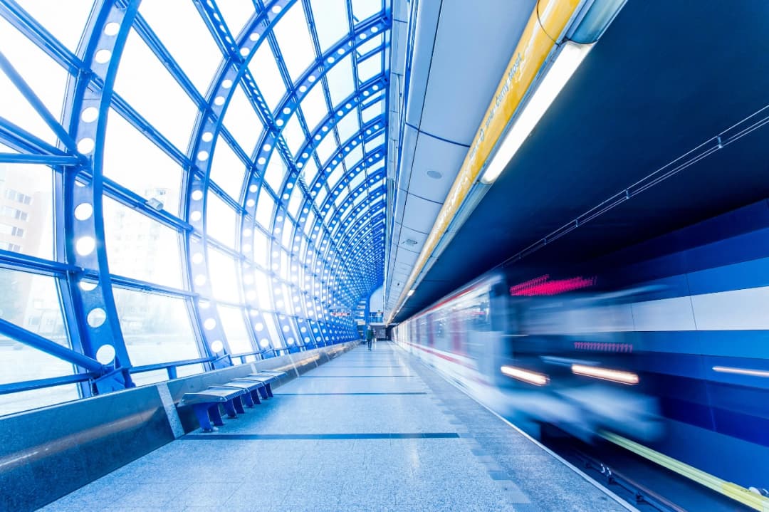 Modern train station platform with a sleek, blue arched design, illuminated by natural light, as a train speeds by, leaving a motion blur effect.