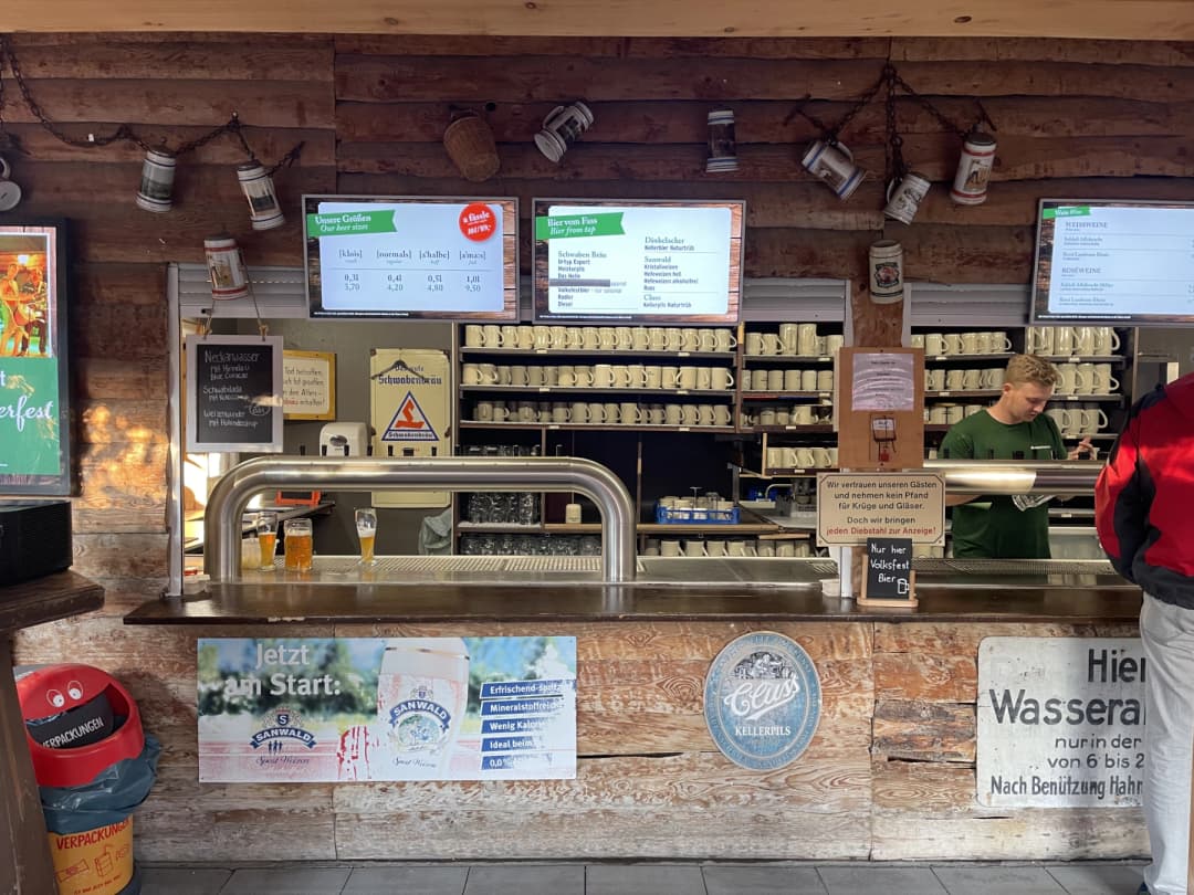 A beer counter at Schwabengarten beer garden, featuring large shelves filled with beer steins, digital menus displaying beer options, and a bartender serving drinks. The rustic decor includes wood paneling and beer advertisements.