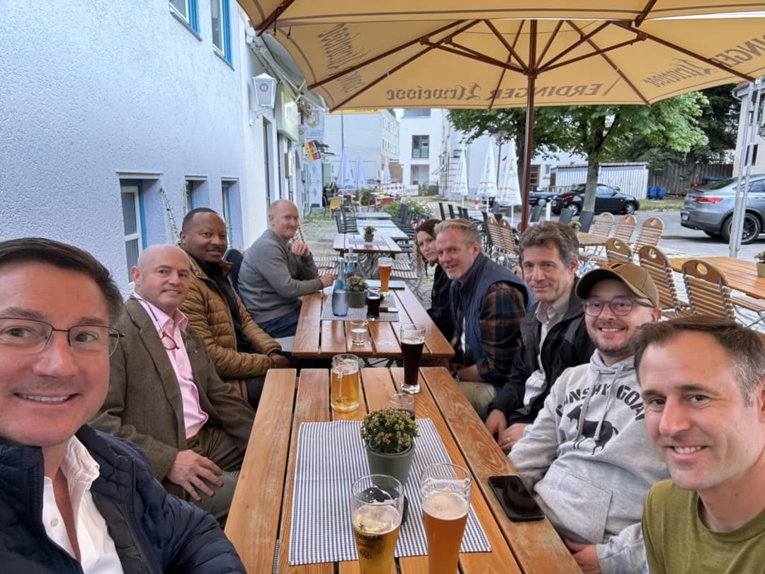 A group of nine people sitting at an outdoor restaurant table under a large umbrella, enjoying drinks and smiling together. The atmosphere is relaxed, with pints of beer on the table, and they are seated in a cozy dining area with wooden benches.