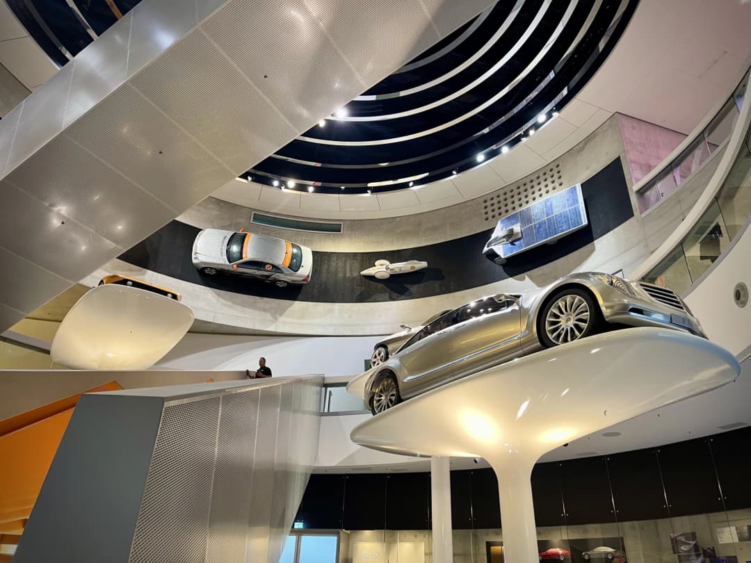 The interior of the Mercedes-Benz Museum in Stuttgart, featuring a sleek, modern design with various cars displayed on platforms and walls. A spiral walkway leads visitors around the exhibits, showcasing the evolution of Mercedes vehicles.