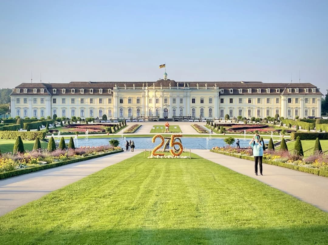 The grand Ludwigsburg Palace with well-manicured gardens and vibrant floral displays, featuring a large '25' made of pumpkins in the foreground as part of the Ludwigsburg Pumpkin Festival celebration.