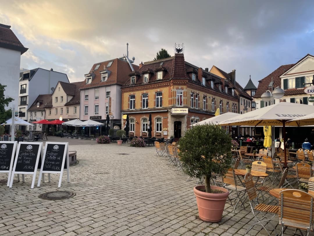A charming town square with traditional European architecture, outdoor seating at restaurants under umbrellas, and cozy street cafes. The sky is partly cloudy with soft light from the setting sun.