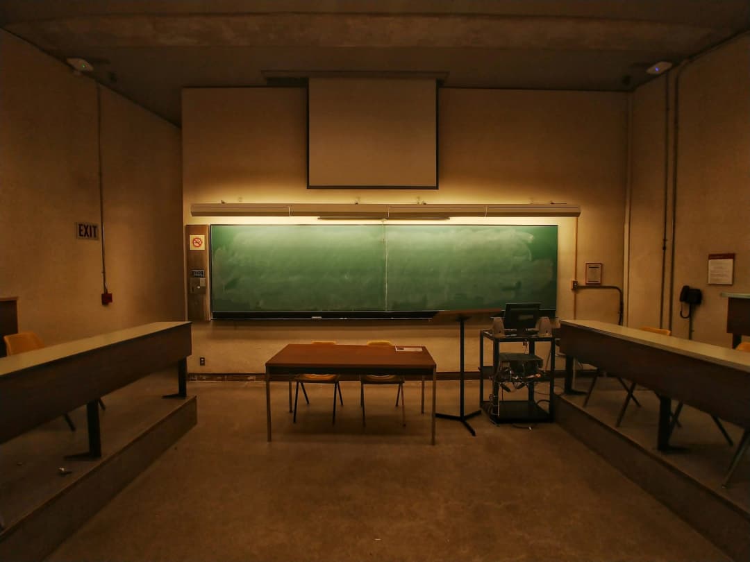 Empty lecture hall with tiered seating, a long green chalkboard, a lowered projector screen, and a teacher's desk in warm dim lighting.
