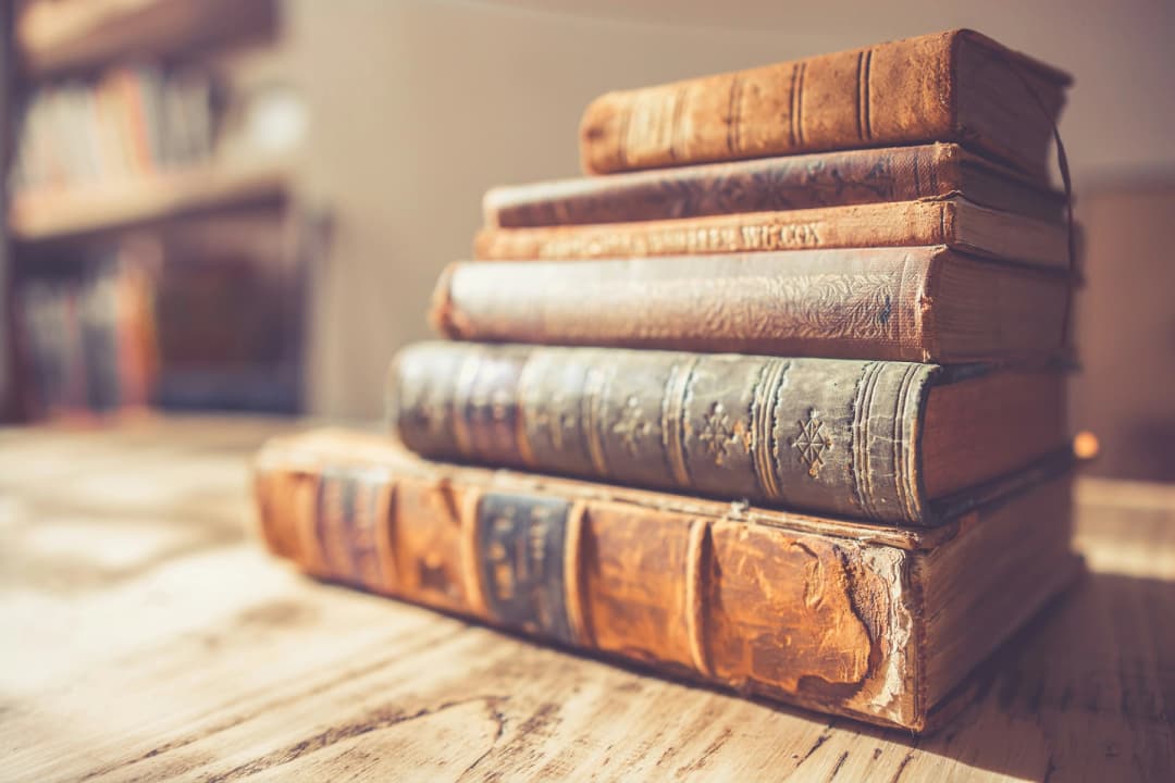 Stack of worn antique books on a wooden table with a blurred bookshelf in the background.