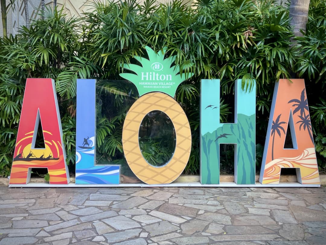 Colorful ALOHA sign with vibrant tropical-themed letters, featuring depictions of sunsets, waves, pineapples, lush green mountains, and palm trees, displayed at the Hilton Hawaiian Village in Waikiki, surrounded by lush greenery.