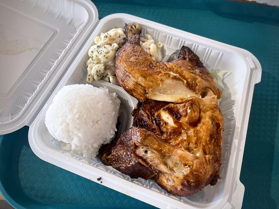 Takeout plate featuring succulent, golden-brown Huli Huli chicken served alongside a scoop of fluffy white rice and creamy macaroni salad, presented on a green tray, showcasing a classic Hawaiian comfort meal.