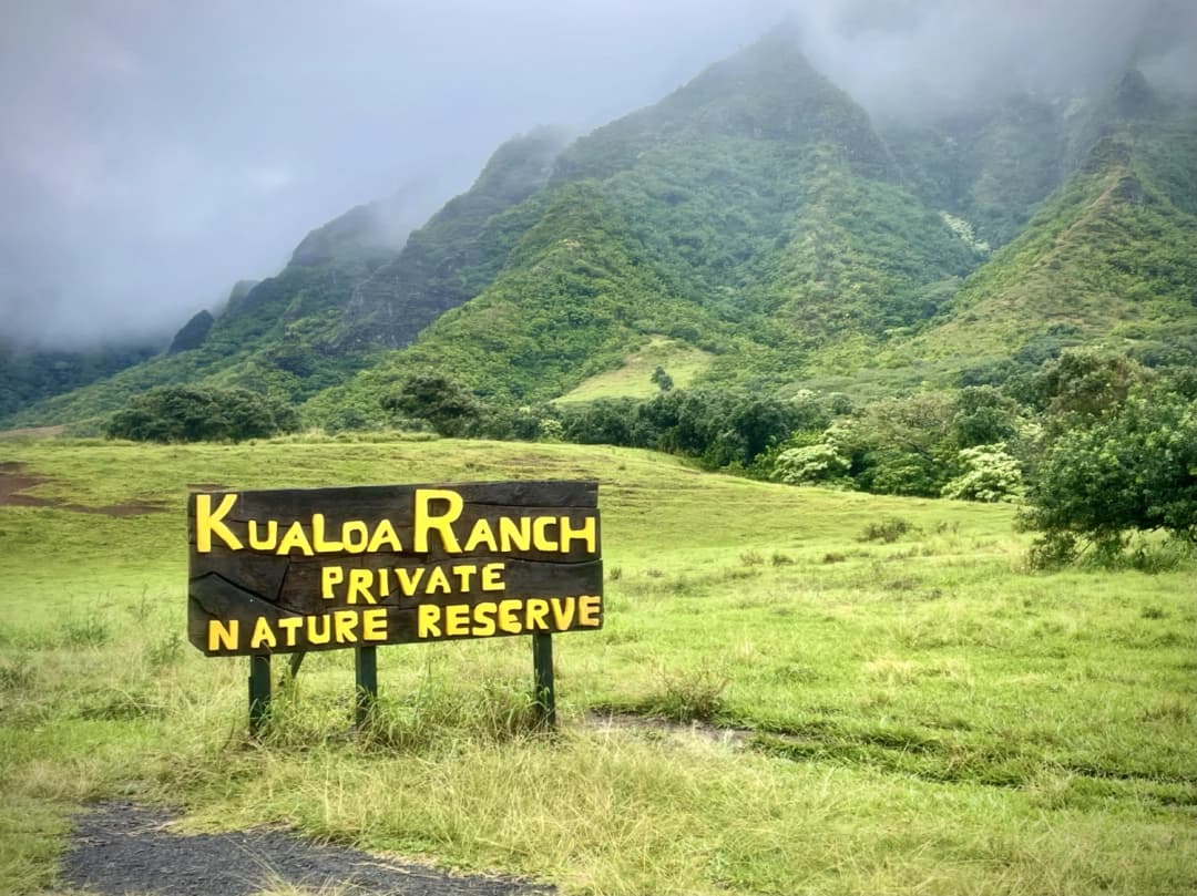 Wooden sign reading 'Kualoa Ranch Private Nature Reserve' set against the lush green backdrop of towering mountains shrouded in mist, capturing the dramatic beauty and tranquility of this iconic Oahu destination.