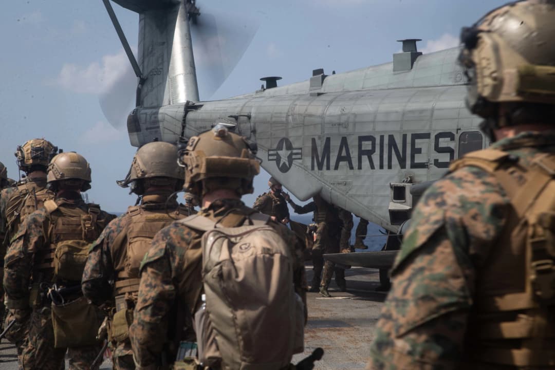 U.S. Marines in full combat gear prepare to board an MV-22 Osprey aircraft, with the word 'MARINES' prominently displayed on the side of the aircraft, symbolizing readiness and operational precision.