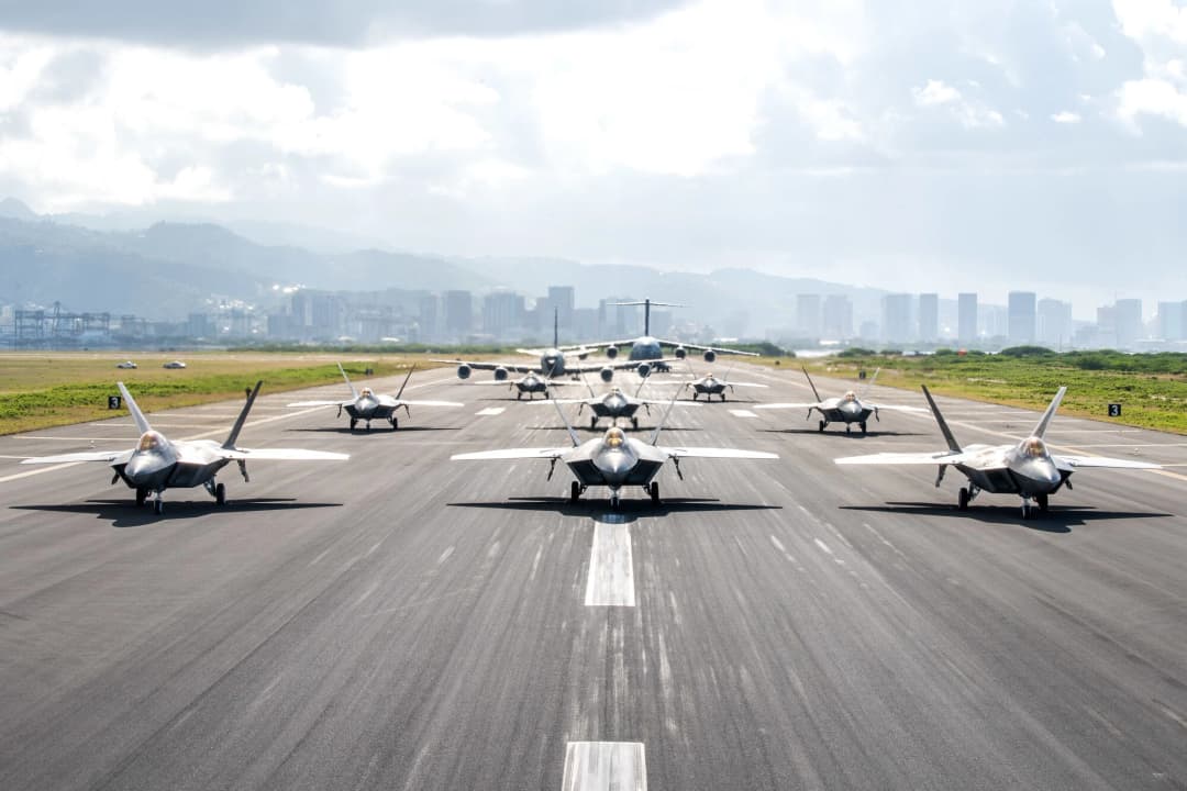 A striking display of military airpower as a formation of F-22 Raptors, supported by additional aircraft, taxi down a runway with the Honolulu skyline and mountains in the background, showcasing readiness and precision in the Indo-Pacific region.