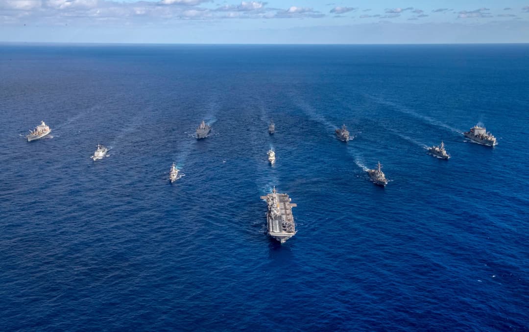 A powerful formation of U.S. Navy vessels, led by an aircraft carrier, sails through the vast blue expanse of the Pacific Ocean under a clear sky, showcasing maritime strength and coordination in the Indo-Pacific region.