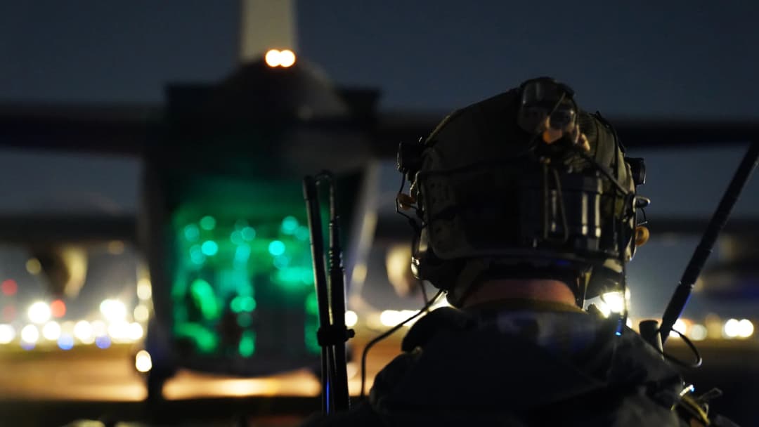 A Special Operations Forces operator, silhouetted against the glowing green interior of an aircraft at night, prepares for a mission, embodying precision and readiness in low-light conditions.