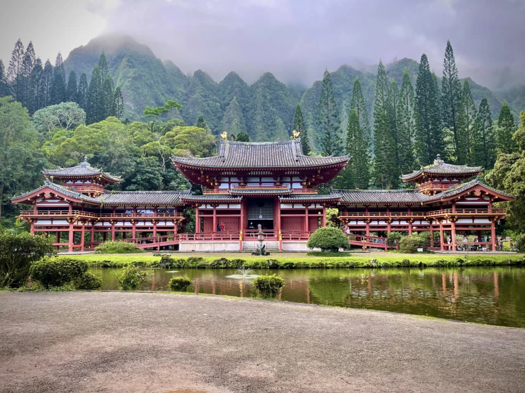 The Byodo-In Temple, a striking red and gold replica of its Japanese counterpart, set against a lush green backdrop of towering Koʻolau Mountains and misty clouds, reflected in the serene koi pond below.