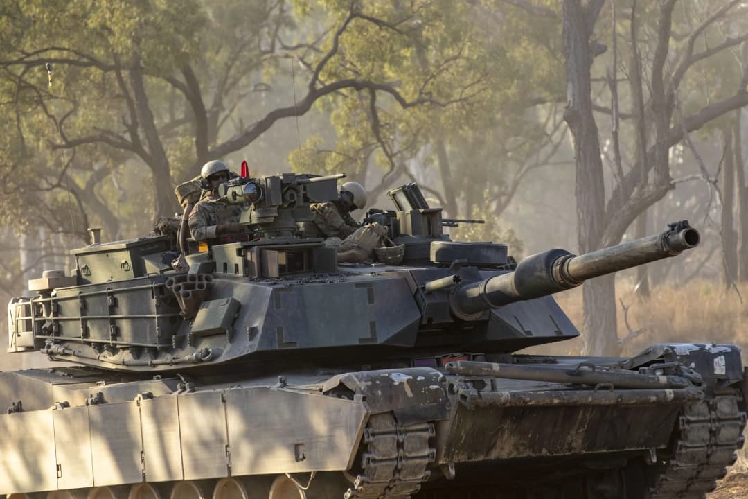 An M1 Abrams tank, crewed by U.S. Army soldiers, maneuvers through a wooded training area under dappled sunlight, showcasing the power and readiness of ground forces in challenging environments.