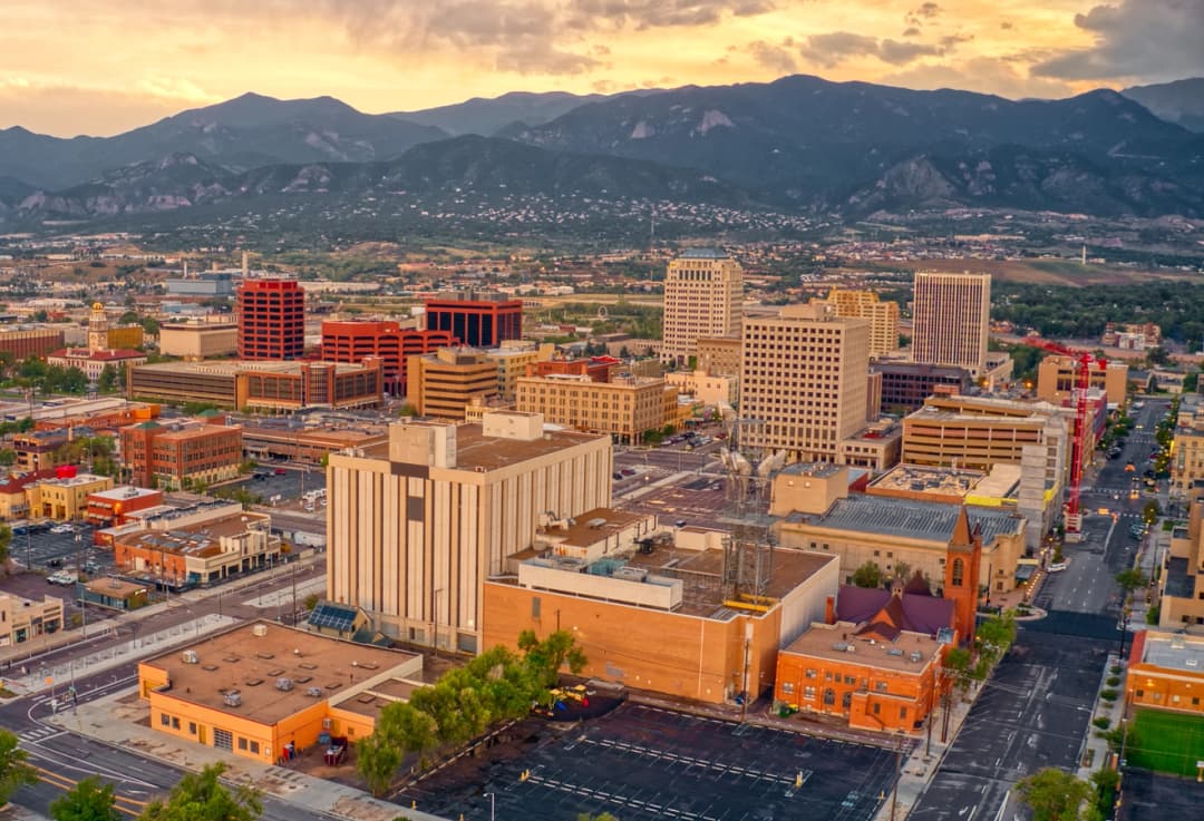 Aerial view of downtown Colorado Springs at sunset, with modern buildings set against a backdrop of the Rocky Mountains and a glowing sky.