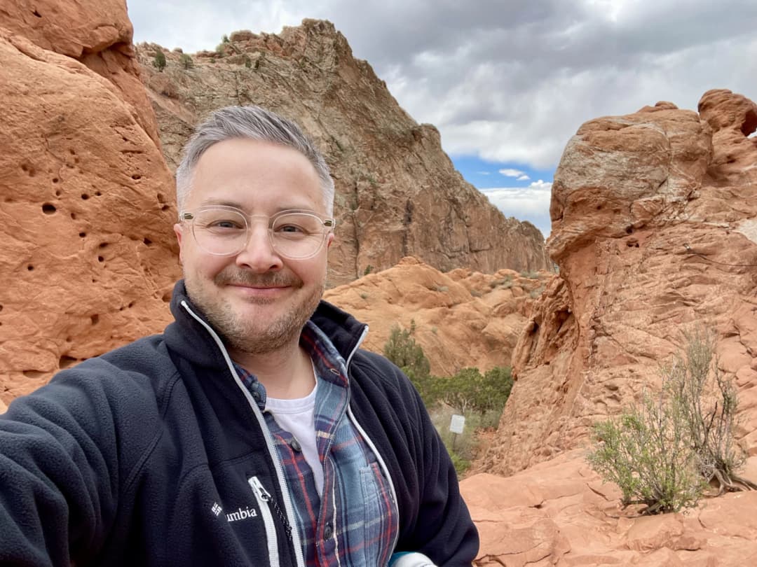 Jeff Thomas takes a selfie at Garden of the Gods, surrounded by towering red rock formations under a partly cloudy sky.