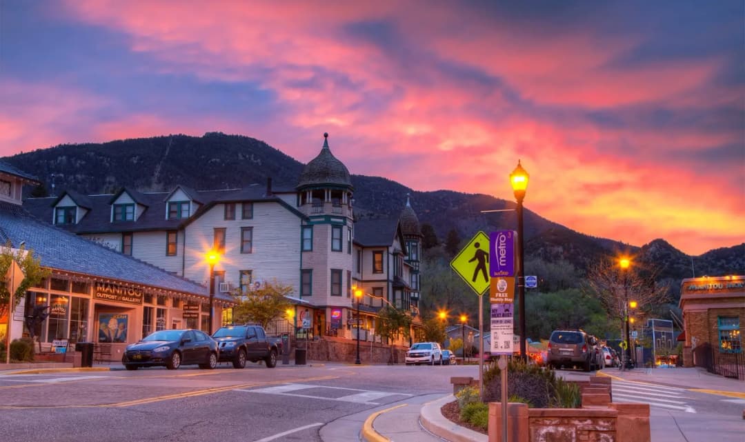 A vibrant sunset paints the sky over historic downtown Manitou Springs, Colorado, with Victorian-style buildings, streetlamps glowing, and cars parked along the street beneath the surrounding mountain landscape.