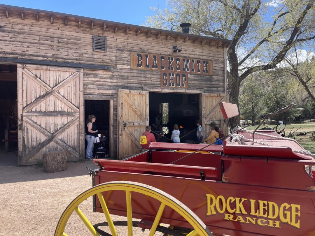 Visitors gather outside the Blacksmith Shop at Rock Ledge Ranch Historic Site, with a bright red and yellow wagon in the foreground and a rustic wooden building set against a clear blue sky and leafy trees.