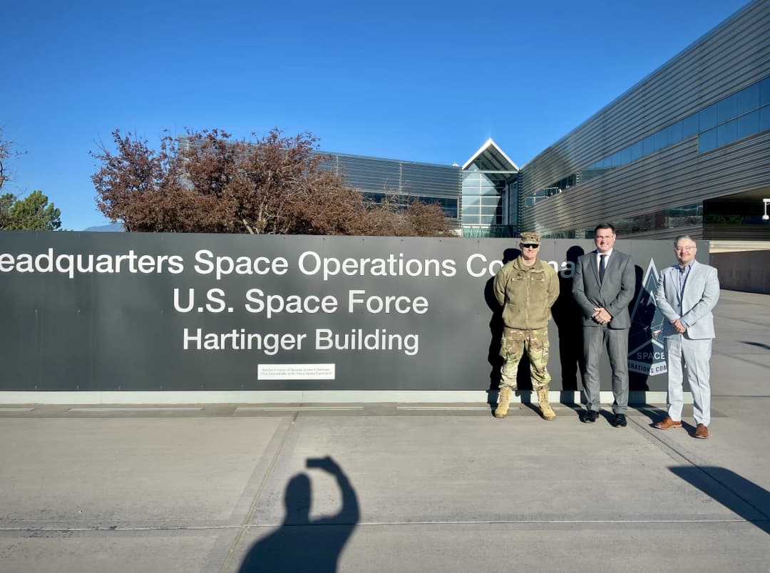 Three individuals—one in military uniform and two in suits—stand in front of the 'Headquarters Space Operations Command, U.S. Space Force, Hartinger Building' sign, with the modern facility and a clear blue sky in the background. A photographer's shadow is visible in the foreground.