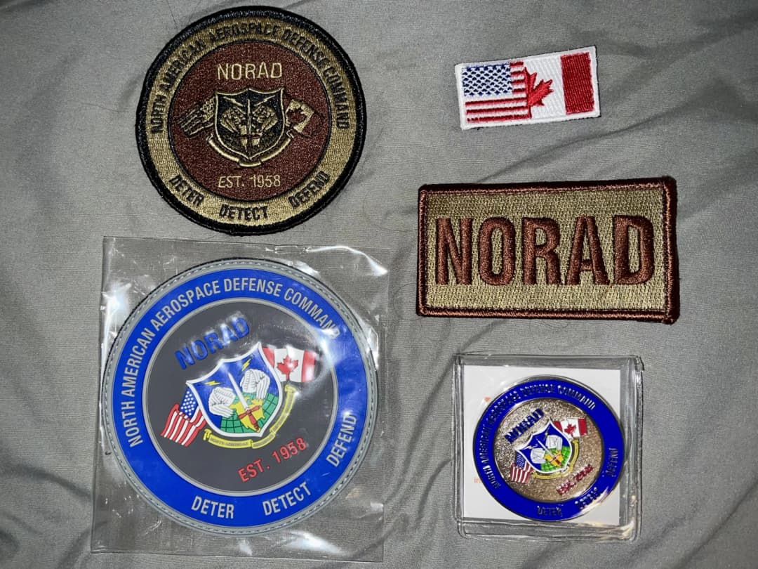 A collection of NORAD-themed swag displayed on a gray surface, including patches with U.S. and Canadian flags, a NORAD emblem coin, and a commemorative medallion with the command's 'Deter, Detect, Defend' motto.