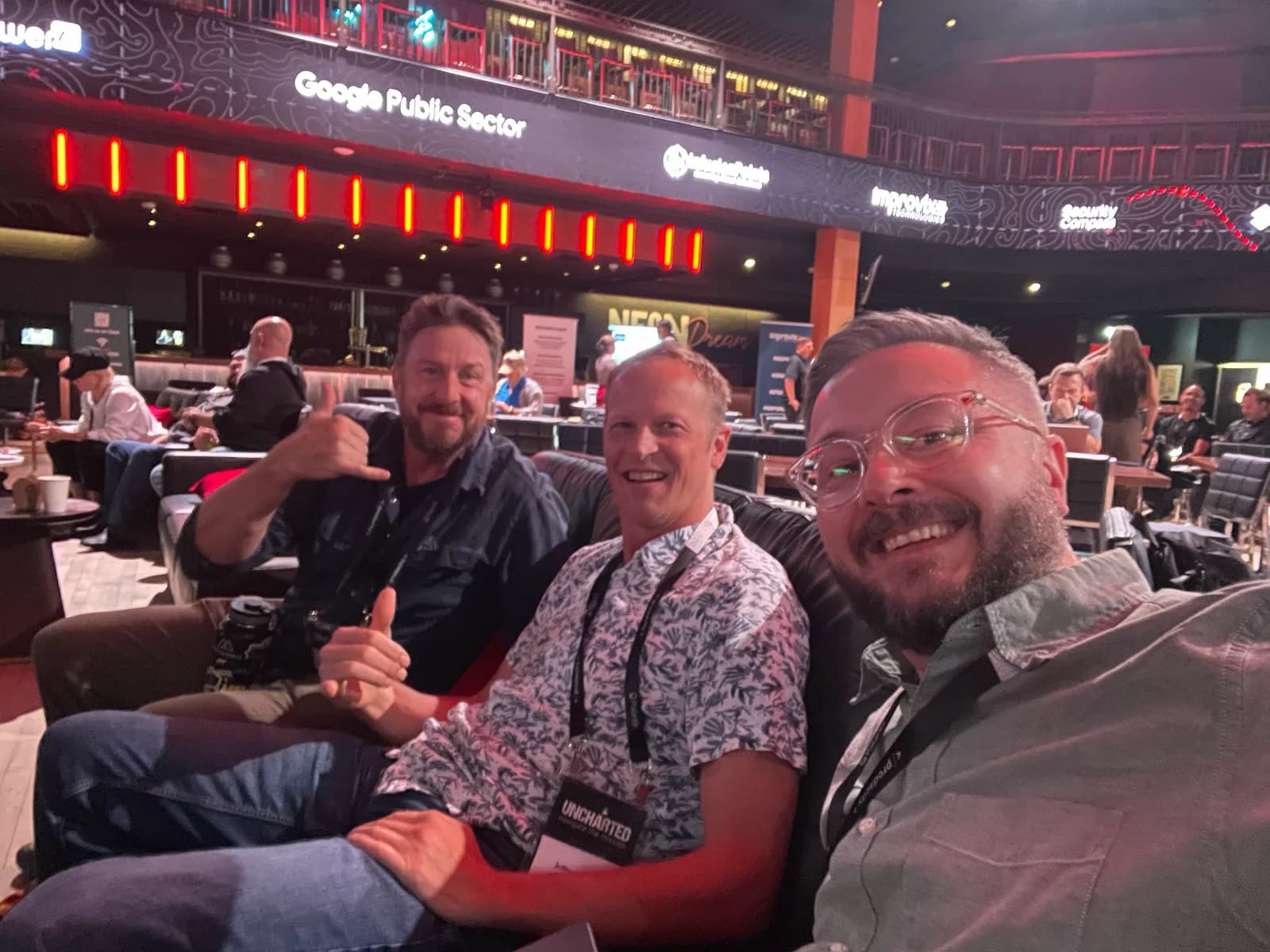 Three conference attendees seated in the venue, smiling at the camera, with Google Public Sector signage on the balcony behind them.