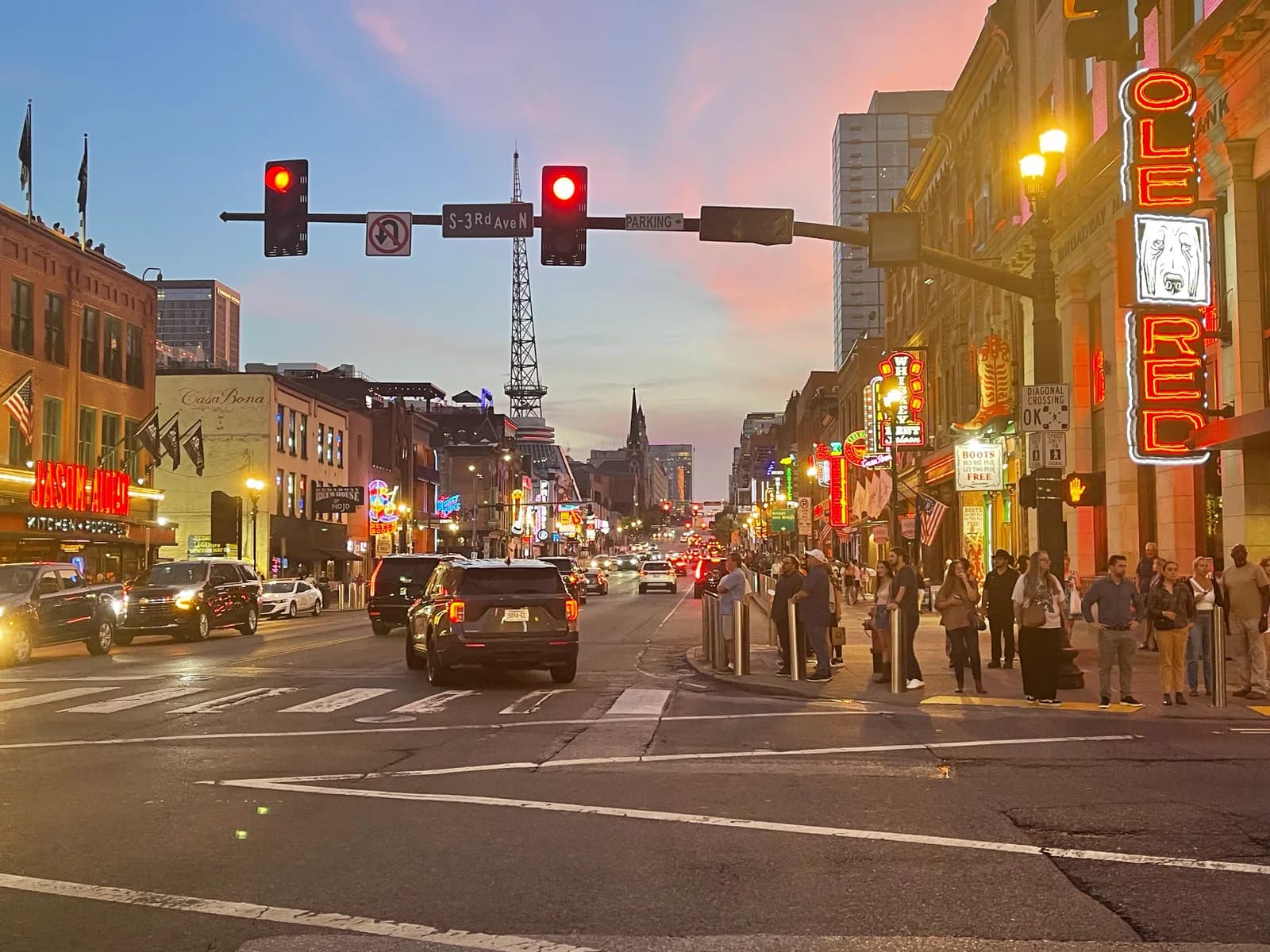 Lower Broadway in Nashville at dusk, neon signs lit, traffic and people at the crosswalk under a pink sky.