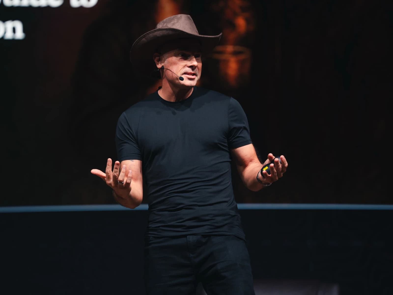 Russ Miles on stage in a cowboy hat and black t-shirt, hands open mid-sentence, his title slide blurred behind him.