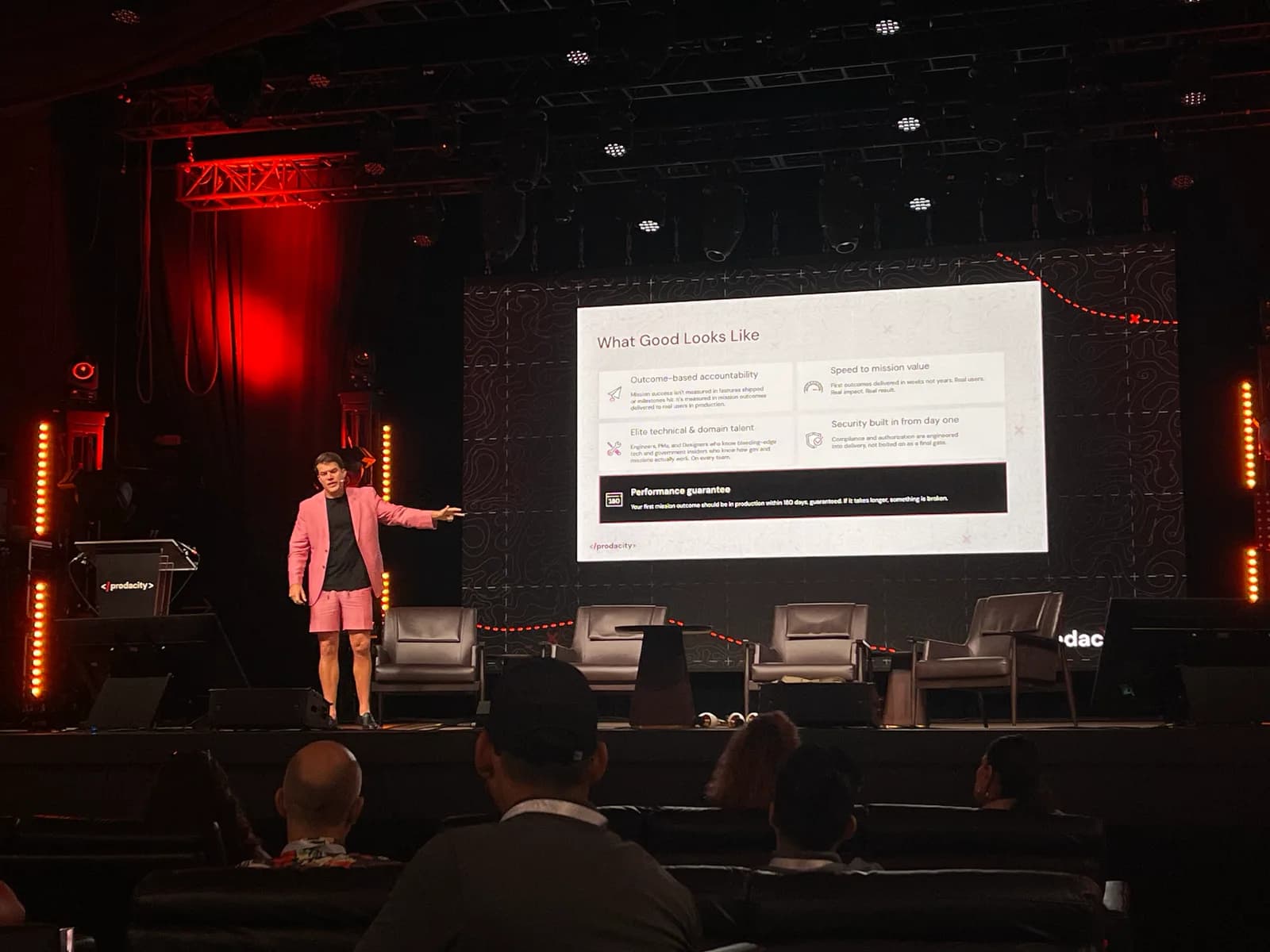 A speaker in a pink jacket gesturing at a slide headed What Good Looks Like, listing outcome-based accountability, speed to mission value and a performance guarantee.