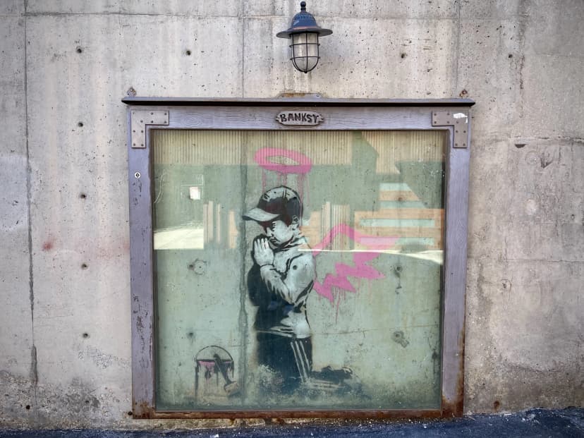 Banksy mural of a kneeling child with a pink halo behind glass on a concrete wall.