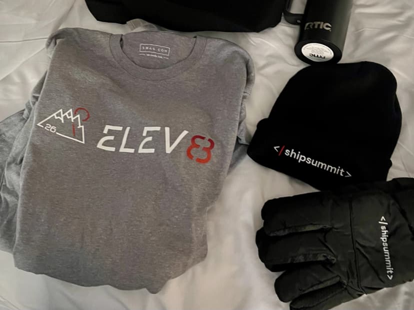 Event swag laid out on a bed: Elev8 shirt, ShipSummit beanie, gloves, and bottle.